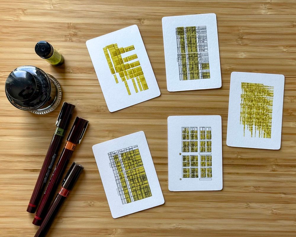 A bamboo desk with five small works of art on them, each 63mm x 88mm. They are in black and yellow, and the artwork is abstract line work based on a recursive algorithm that subdivides space in a regular way. The results are reminiscent of technical drawings. There are three Rotring Isograph pens on the left side of the trading cards, and two bottles of ink above them.