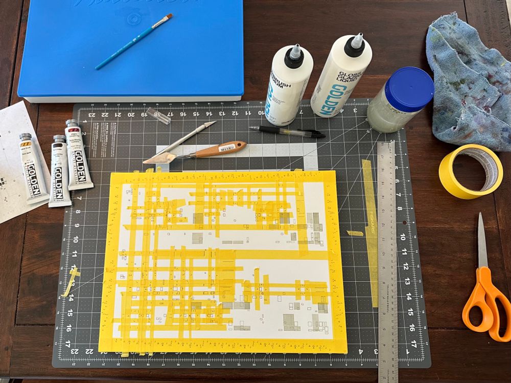 An in-progress photo of a painting in which yellow masking tape is being used to block out sections that will be painted. There are a variety of other tools scattered on the table around it, including scissors, a ruler, and various tubes of acrylic paint and bottles of medium. 