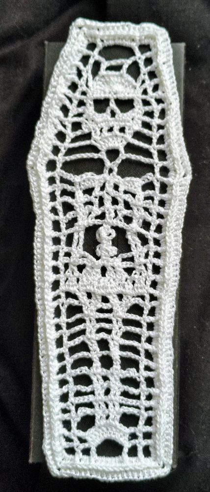 A white crochet bookmark. Pattern is a skeleton lying in a coffin.