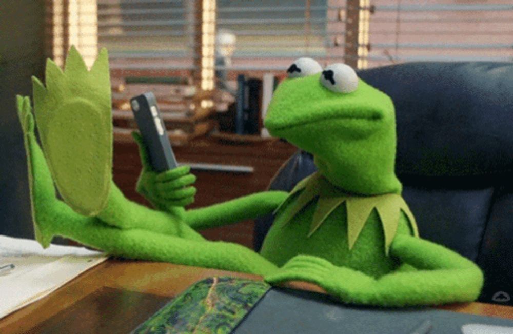 kermit the frog is sitting at a desk with his feet up holding a cell phone