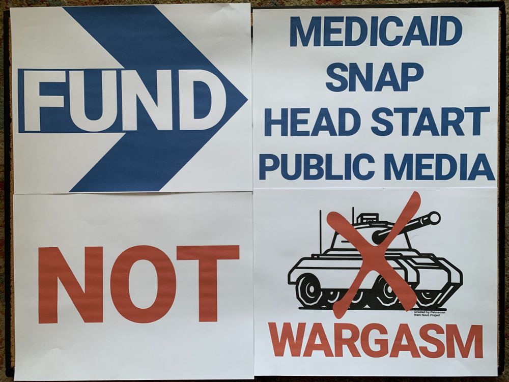 A photo of a sign made of four panels. The top left panel has a thick blue arrow with the word FUND in white. The top right panel has the words MEDICAID, SNAP, HEAD START and PUBLIC MEDIA in blue. Lower left panel says NOT in red. Lower right panel depicts a drawing of a tank with a red X across it and the word WARGASM at the bottom.