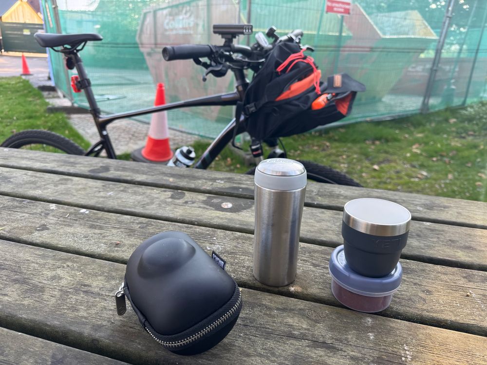 A Giant Roam 0 leaning against a picnic table in a park. On the table is a Wacaco Picoexpress portable espresso maker, a small stainless steel thermo flask, a Yeti Espresso cup, and a small pot of ground coffee. 

