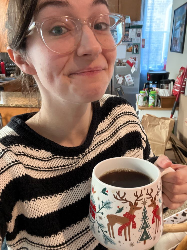 Claire, holding a festive holiday mug of coffee, where’s a striped black and white sweater and her hair pulled back, with clear glasses and a smile in this selfie