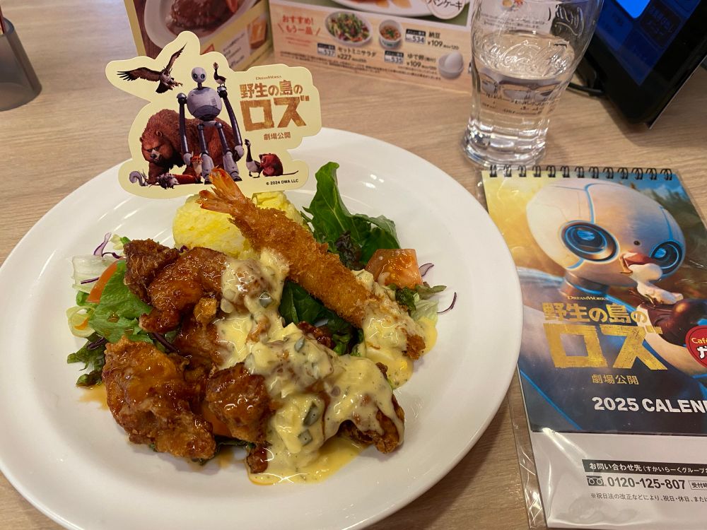 テーブルの上の写真です。手前にある白い皿の上にはチキン南蛮、エビフライ、黄色いライス、サラダが乗っています。ライスには「野生の島のロズ」のキャラクターが描かれた紙が刺さっています。皿の右隣には「野生の島のロズ」の卓上カレンダーがあります。奥には水の入ったグラスがあります。