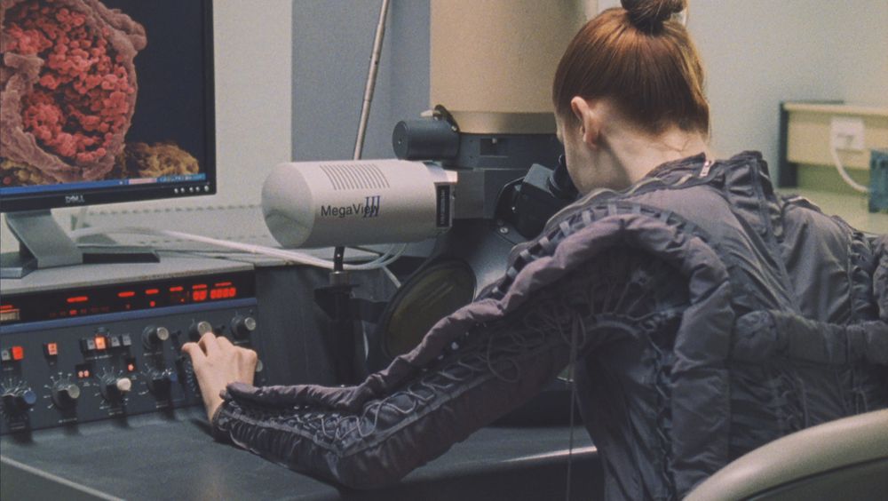 A film still showing a person with red hair and dark top from behind, as they sit at a scientific desk examining something through a viewfinder