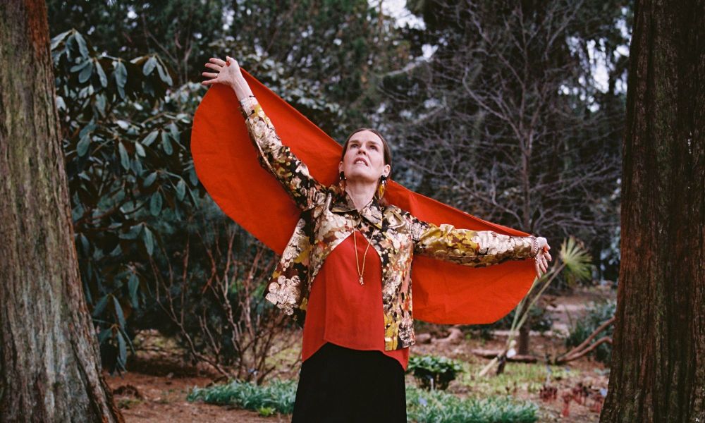 A middle aged woman in a red cape holds her arms apart and looks at the sky, stood in a clearing in a wood