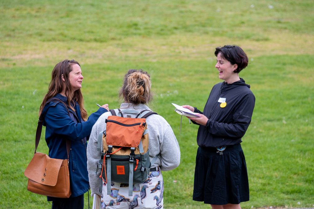 Three people gathered in a circle on a patch of green grass, laughing and talking in outdoor clothes and bags