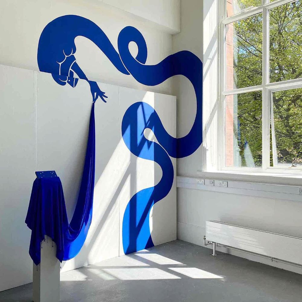 photograph of a white walled exhibition space with a sunny window looking out onto a tree on the right. on the left, a blue graphic hand swirls up the side, holding on to a blue cloth that drapes over a plinth in the centre. 