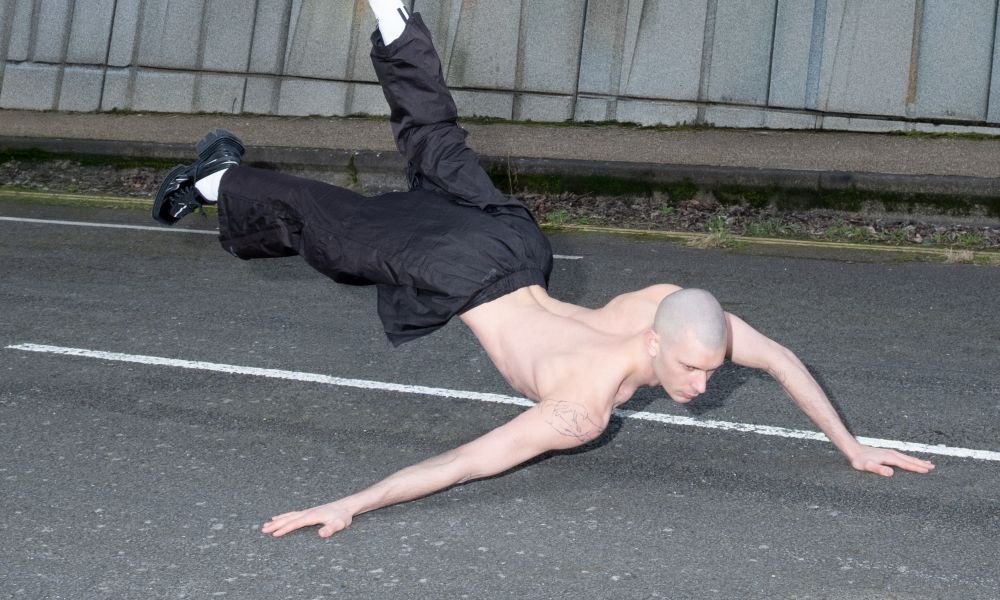 A white shirtless person with shaved head in tracksuit bottoms positions their legs in the air with hands on a road