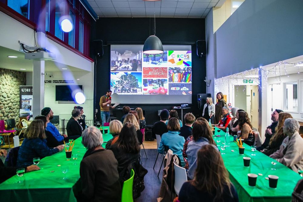 A group of people sit on two long green tables looking at someone giving a presentation on a large screen at the end of the room