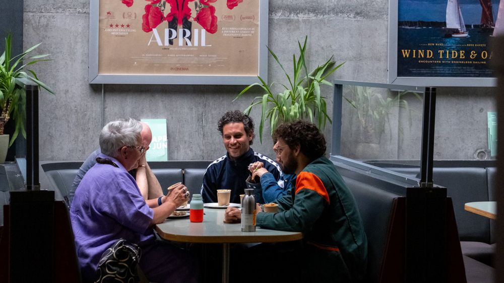 Four people sat around a booth table in a cafe, smiling and chatting, posters on the wall and plants on the side