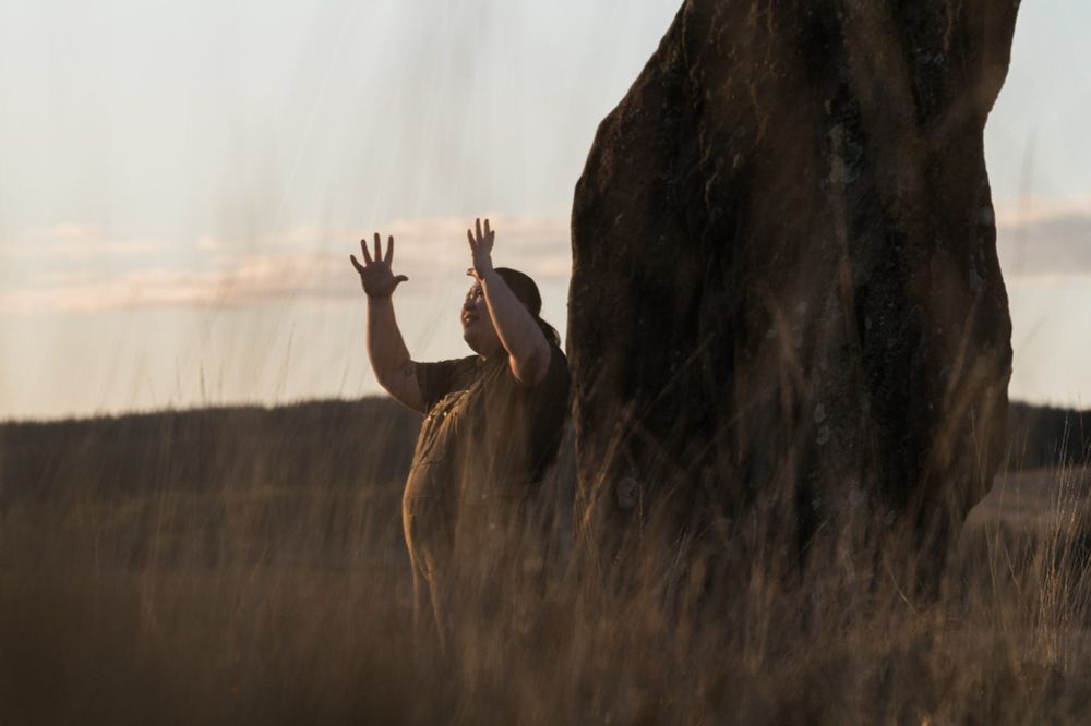 A person holds arms above them, stood next to a large boulder in a wheatfield