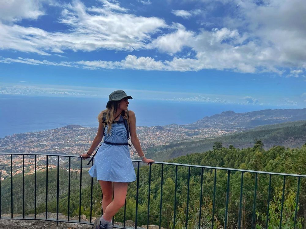 Girl in blue summer dress with a hiking hat, rucksack and hiking boots standing looking to her left, leaning against a railing at a view point, with a view of the sea and city of Madeira behind