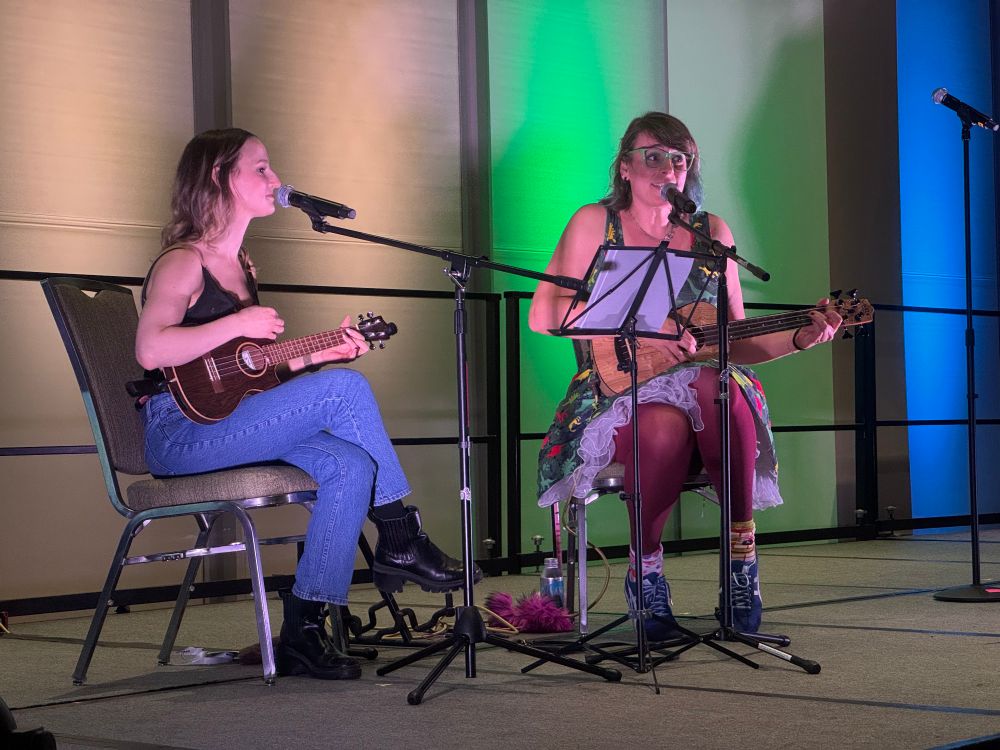Two ukulele players on a stage! On the left, jackie is holding a tenor ukulele and is singing in a mic. On the right, Valerie with a U-bass, singing and looking nervously excited 
