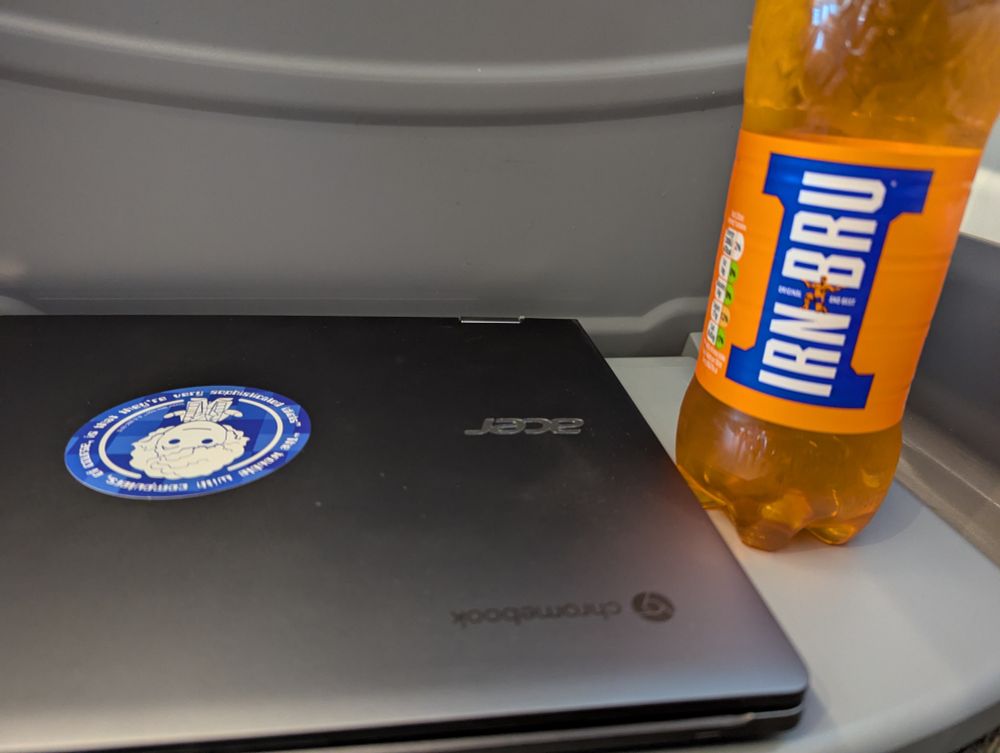 A closed laptop and a plastic bottle of Irn Brun on a train seat table.