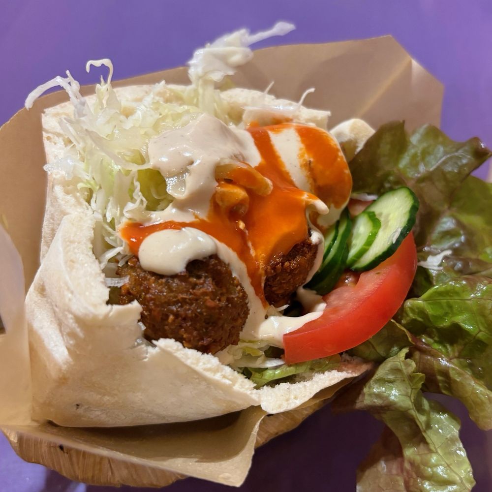 オープンピタサンドにトマト、きゅうり、レタス、キャベツ、ファラフェルが入っていて、上から白とオレンジのドレッシングがかかっている。サンドはベージュの包み紙に入っている。背景は紫色。