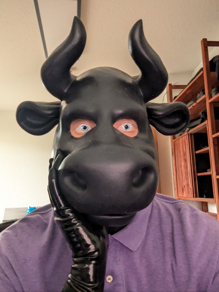 A silicone cow mask with horns stares off-center at his work, his silicone eyes deep in thought. He wears a purple polo shirt. His head is resting on one of his shiny rubber hands.