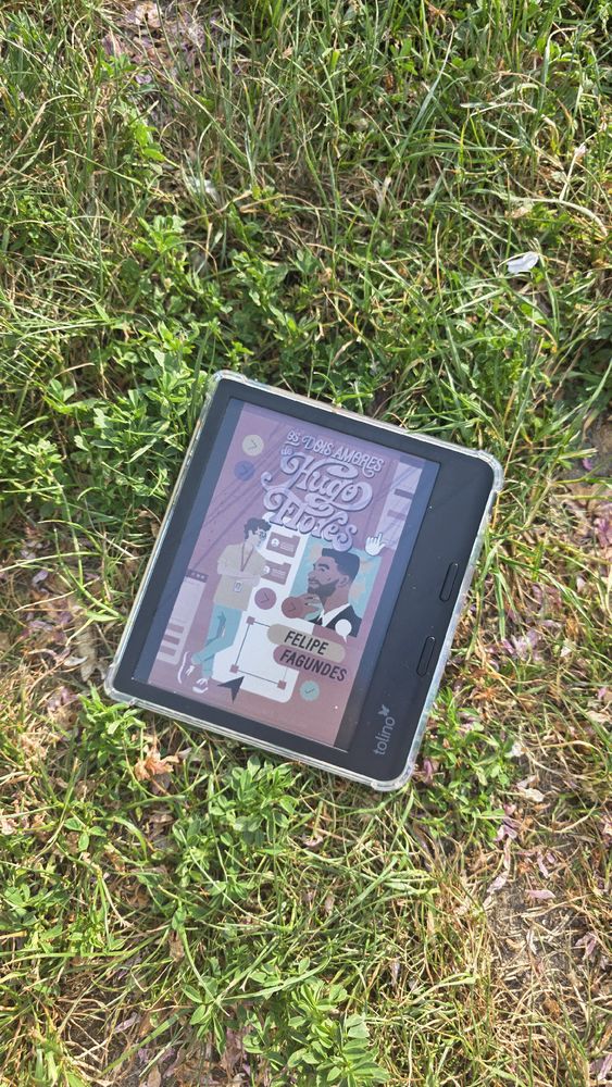 Um e-reader da marca Tolino está apoiado na grama, com a tela exibindo a capa do livro Os Dois Amores de Hugo Flores, de Felipe Fagundes. A ilustração da capa mostra dois homens: um com pele clara, óculos e crachá, em pé ao lado de telas e ícones de interface digital; o outro com pele escura, barba e camisa social, olhando com expressão séria. O fundo é rosa com detalhes em lilás, vermelho e azul, com elementos gráficos que remetem ao universo da tecnologia e design. O aparelho está em uma capa transparente e a grama ao redor tem tons de verde e roxo, com folhas secas espalhadas.