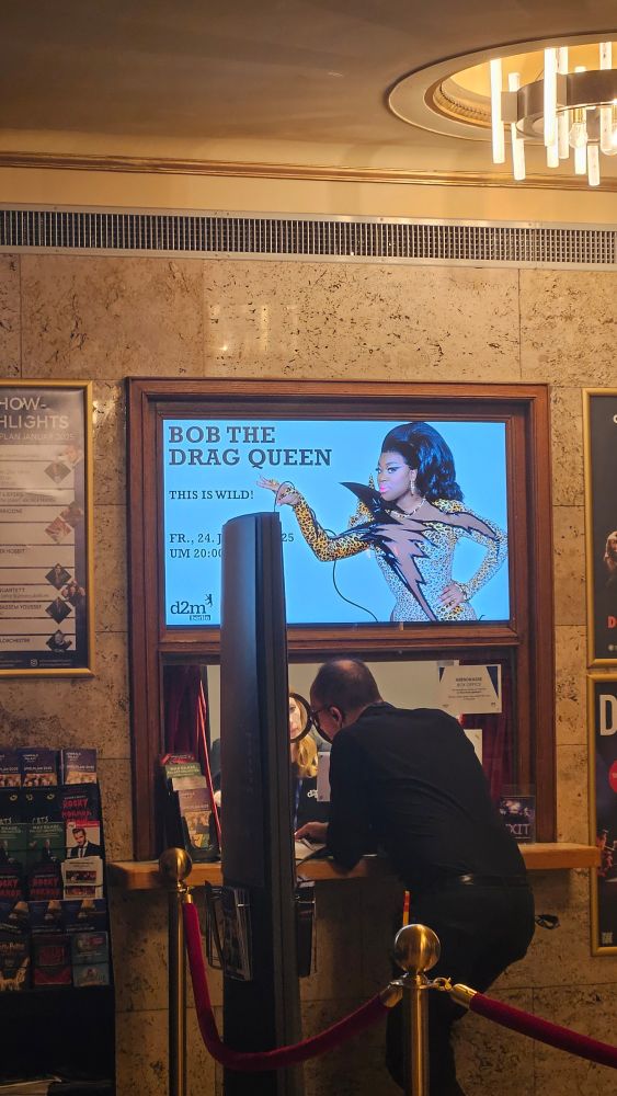 Foto tirada no interior de um teatro, com uma bilheteria em destaque. Na parte superior, há um cartaz digital promovendo o show de "Bob the Drag Queen" com o título "This Is Wild!" e a data "24 de janeiro de 2025, às 20h". Abaixo, um atendente está auxiliando um cliente no balcão, com cordões de isolamento vermelho delimitando o espaço.