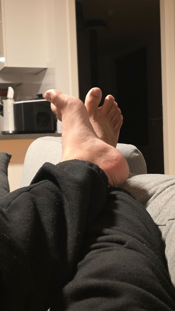 foto dos meus pés entrelaçados. calça preta. em cima do encosto do sofá cinza. atrás dos pés, tem o restante da casa: un cômodo aberto escuro e um pedaço de uma cozinha americana.