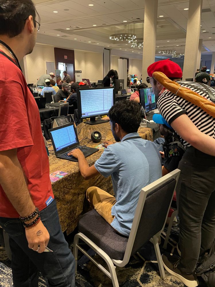 3 or 4 con attendees hover over a laptop to solve a puzzle. One of them is in a black-and-white striped shirt, a red beret, and has a baguette slung over his back. 