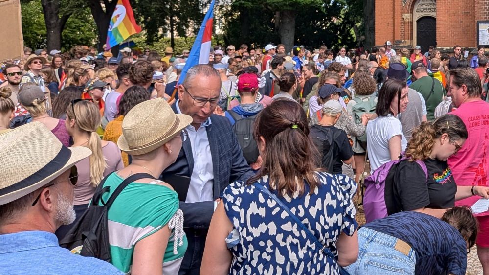 Menschenmenge beim CSD Bautzen vor einer Kirche, viele tragen bunte Kleidung und Regenbogenflaggen. Im Vordergrund spricht der Bürgermeister von Bautzen im dunklen Anzug mit Teilnehmenden.