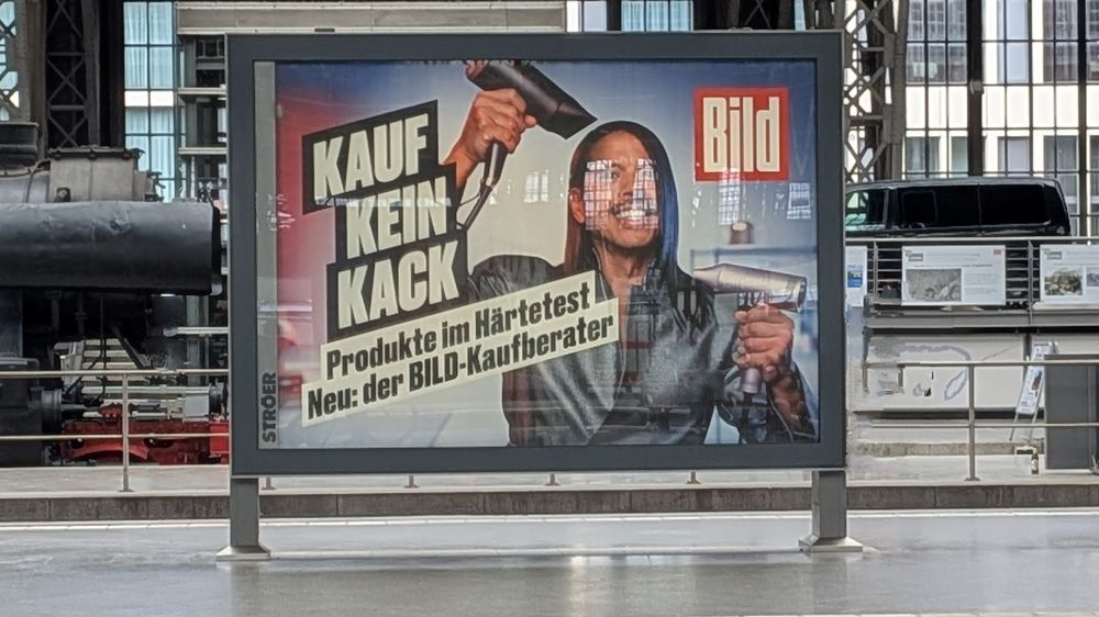 Plakatwerbung am Bahnhof: Eine Person hält in jeder Hand einen Fön und lächelt breit. Daneben der groß gedruckte Slogan „KAUF KEIN KACK“ in gelben Blockbuchstaben. Darunter in kleinerer Schrift: „Produkte im Härtetest – Neu: der BILD-Kaufberater“. Rechts oben ist das rote BILD-Logo zu sehen.