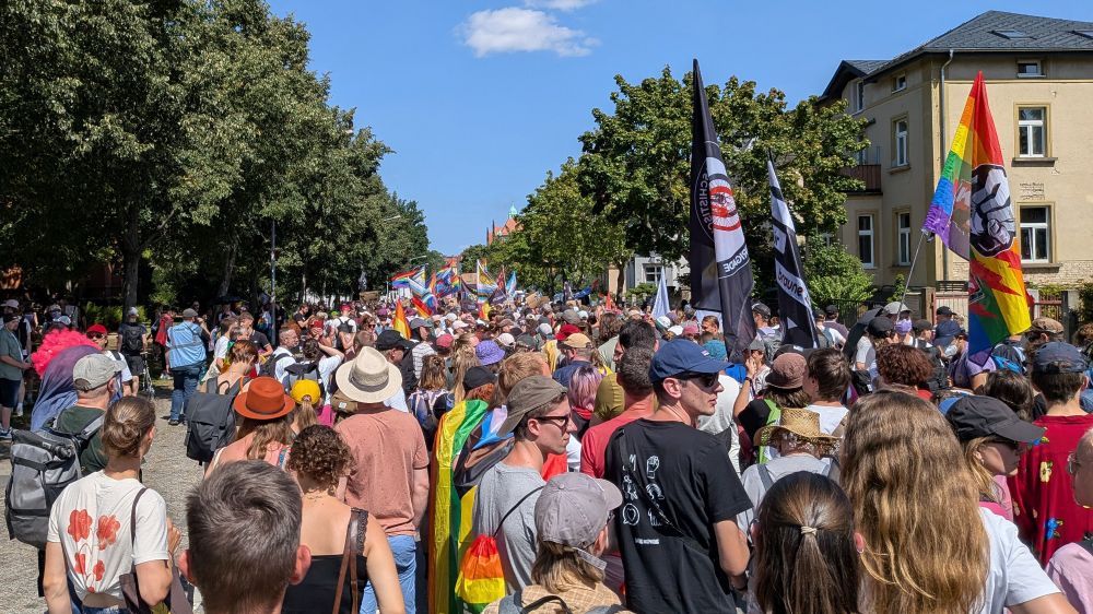 Eine große Menschenmenge nimmt an einer Demonstration oder Parade teil, viele tragen bunte Kleidung und Regenbogenfahnen. Mehrere Banner und Fahnen mit politischen Botschaften sind sichtbar. Die Veranstaltung findet an einem sonnigen Tag auf einer von Bäumen gesäumten Straße vor Wohnhäusern statt.