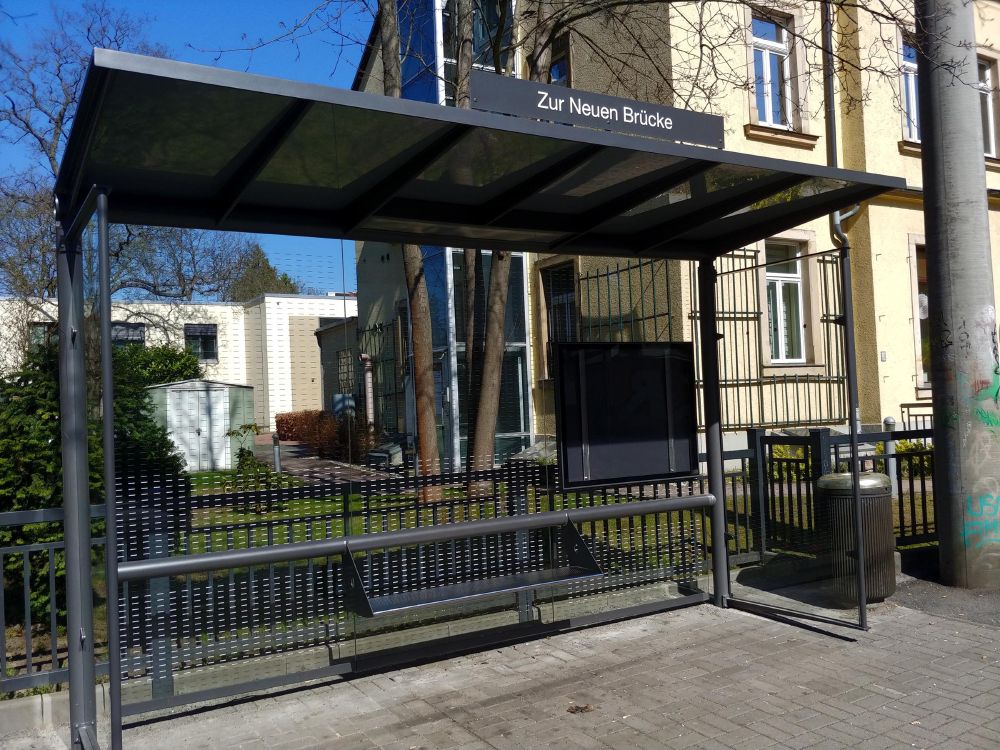 Bushaltestelle mit dem Namen „Zur Neuen Brücke“ bei sonnigem Wetter. Die moderne Haltestelle hat ein dunkles, undurchsichtiges Dach aus Metall oder getöntem Material sowie Glaswände mit Punktemuster. Darunter befindet sich eine schwarze Sitzbank aus gelochtem Metall. Rechts im Bild steht ein Abfalleimer, daneben eine mit Graffiti beschmierte graue Säule. Im Hintergrund sind ältere Gebäude mit gelber und weißer Fassade zu sehen, sowie einige Bäume mit kahlen Ästen. Die Haltestelle ist leer, es sind keine Personen anwesend.
