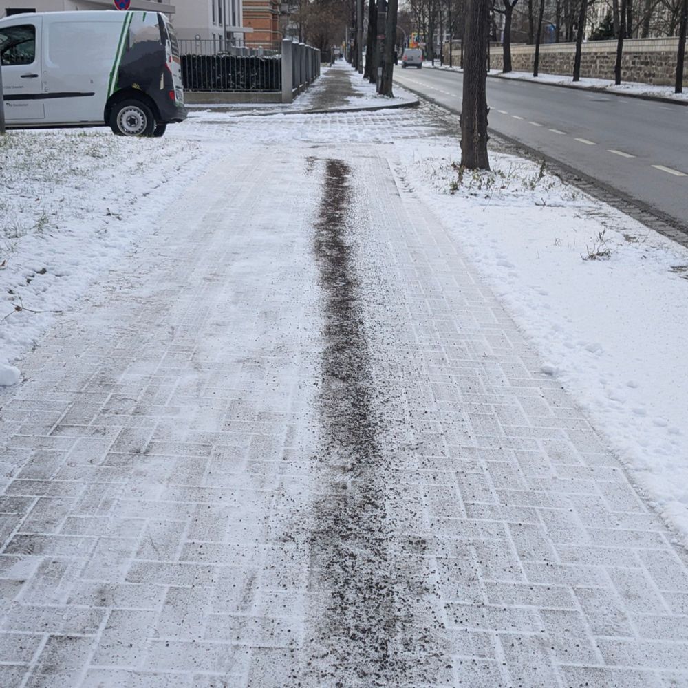 Ein schneebedeckter Gehweg, auf dem eine dünne, unregelmäßige Spur aus Streusand oder Streugut verläuft. Der Weg ist größtenteils ungestreut, was die Sicherheit bei Schnee und Glätte beeinträchtigen könnte. Im Hintergrund sind ein parkender Lieferwagen und eine Straße sichtbar.