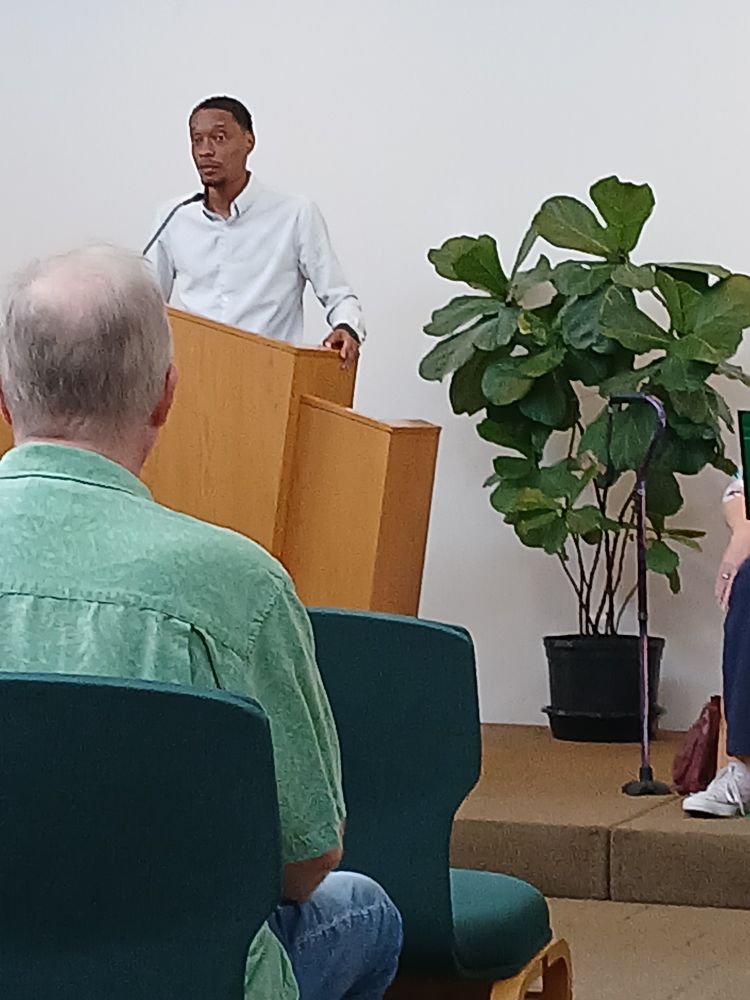 Jordan Bridges director for the New Orleans Worker's Center for Racial Justice. Tall slender Black man with white long sleeve shirt standing at a lectern at the Community Church of Universalist Unitarian Church in New Orleans, LA on fleur de list. He is flanked on the left by a green plant. 