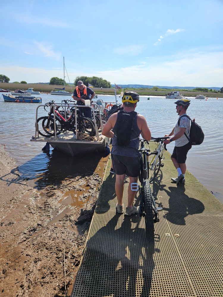 Two bike riders wait on a slipway as bikes are loaded onto ferry. The river is calm and muddy.