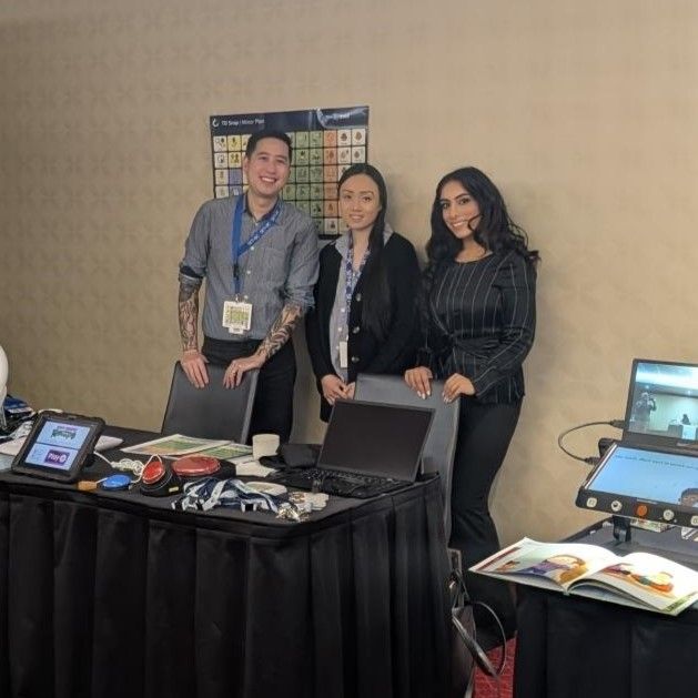 Three people stand behind a table with tech devices and materials at a conference. A poster with a sign language chart is on the wall.