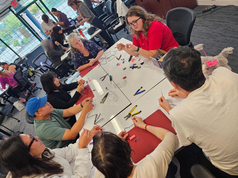 Looking down the length of a large meeting room with desks of people sitting or standing around tables beginning to make items. Visible are matts, plyers, wires.  In the forefront table, one person seems to be instructing others who are concentrating on what she is showing them.