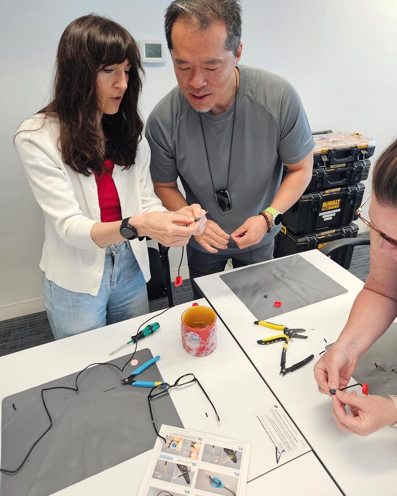 People look at parts together. They stand over a table with mats, tools and a printed page with images and instructions.