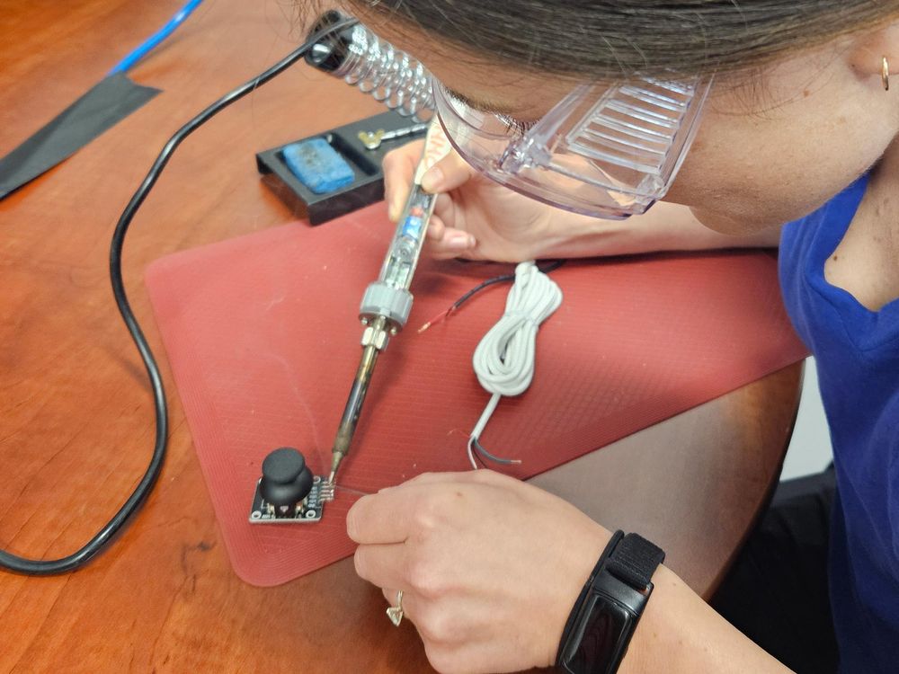 A person wearing eye protection appears to be using a tool to connect wires to a device.