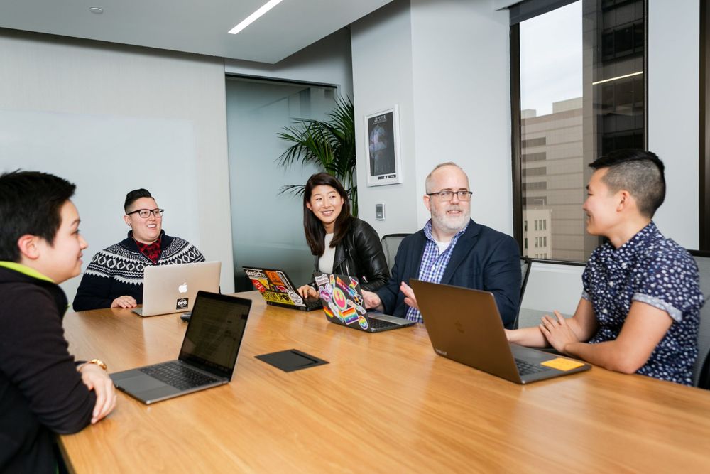 Five people gathered around a conference table in a modern office, engaged in conversation and using laptops. The group is diverse in age, gender, and style, with expressions suggesting engagement and collaboration.