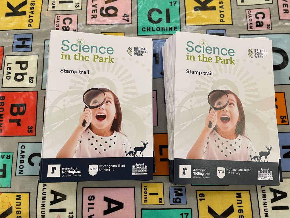 Two piles of leaflets for the science in the Park Stamp trail, set on top of a table cloth with different elements on it (symbol, atomic number etc)
