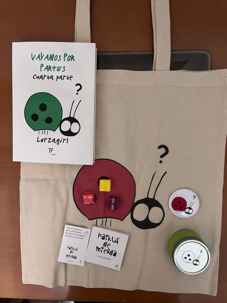 Una bolsa de tela con una mariquita en rojo, con tres August, una chapa con la misma imagen, un bote verde con una vela de manzana, un libro blanco con una mariquita en verde de título “Vayamos por partes cuarta parte”, junto con un pequeño librito de título “Haikus de mierda”.