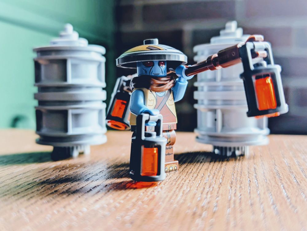 A LEGO Star Wars minifigure of a Duros alien carrying power cells on a beam over his shoulder. The Duros is wearing comfortable clothing, a scarf, and a broad hat.