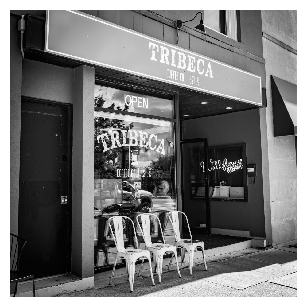 Panchromatic picture of 3 empty chairs outside the Tribeca coffee shop and a sole figure sitting inside by the front window of the shop