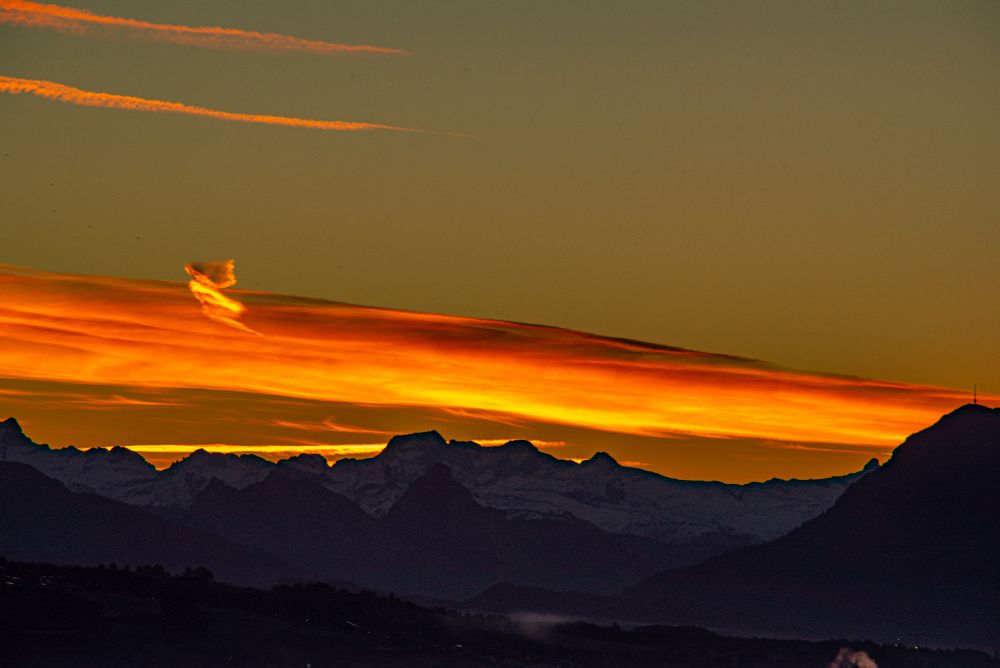 Morgenstimmungen in den Bergen. Die Bergwelt liegt noch mehrheitlich im Dunkeln, einzelne Konturen sind aber schon auszumachen. Ein Wolkenband zieht sich von links nach rechts. Es ist von der, noch nicht sichtbaren Sonne, kräftig gelb-orange eingefärbt. Am oberen Teil des Wolkenbandes, im linken Bildteil, ist eine Wolkenausstülpung sichtbar, die aussieht, wie wenn ein Engel aus dem Wolkenband entfliehen möchte. 