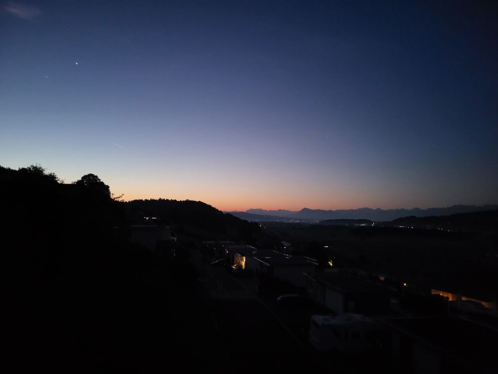 Morgenstimmung mit Venus und Jupiter am wolkenlosen Himmel. Über der Bergkette im Osten ist ein schmales, rosa Band sichtbar. Der Vordergrund liegt noch im dunkeln, ab und an sind Lichter von vereinzelten Häusern erkennbar. 