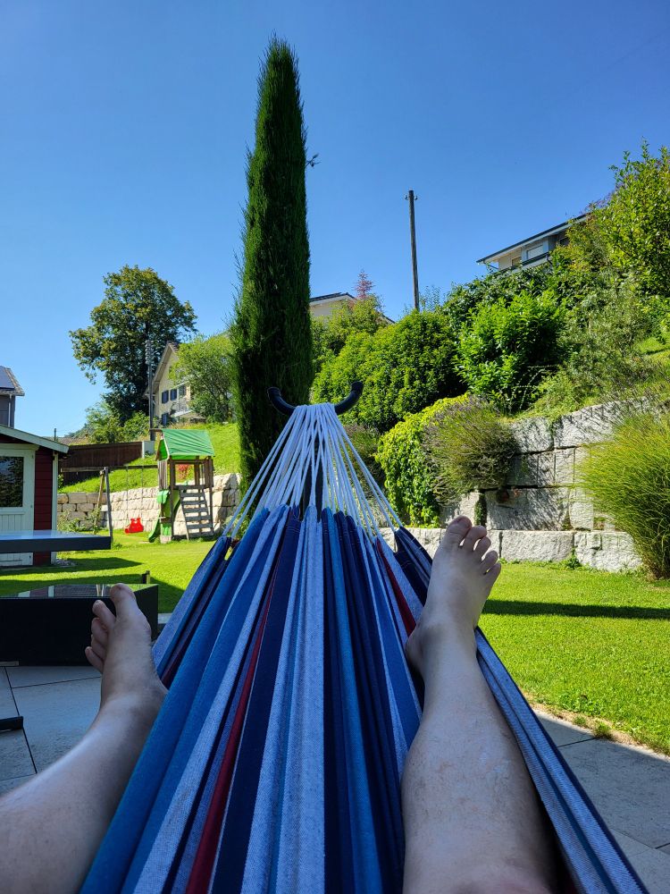 2 Beine in einer Hängematte. Im Hintergrund eine Gartenanlage mit Gartenhäuschen und Spielturm. Und viel blauer Himmel. 