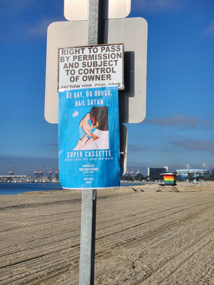 Poster for super cassette saying "be gay do drugs hail Satan" in front of rainbow lifeguard tower in long Beach