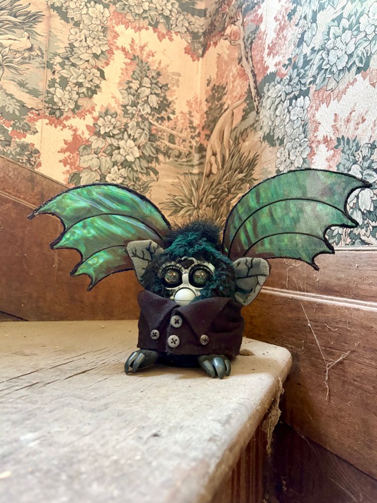 a 1998 Furby that’s been customized to look like Aury from Lily Mayne’s “The Rycke” book. He has green hued black fur, wears a black coat, has gray face, and talons on his toes. Greenish black wings rise up from his back. he sits in a dingy stairwell 