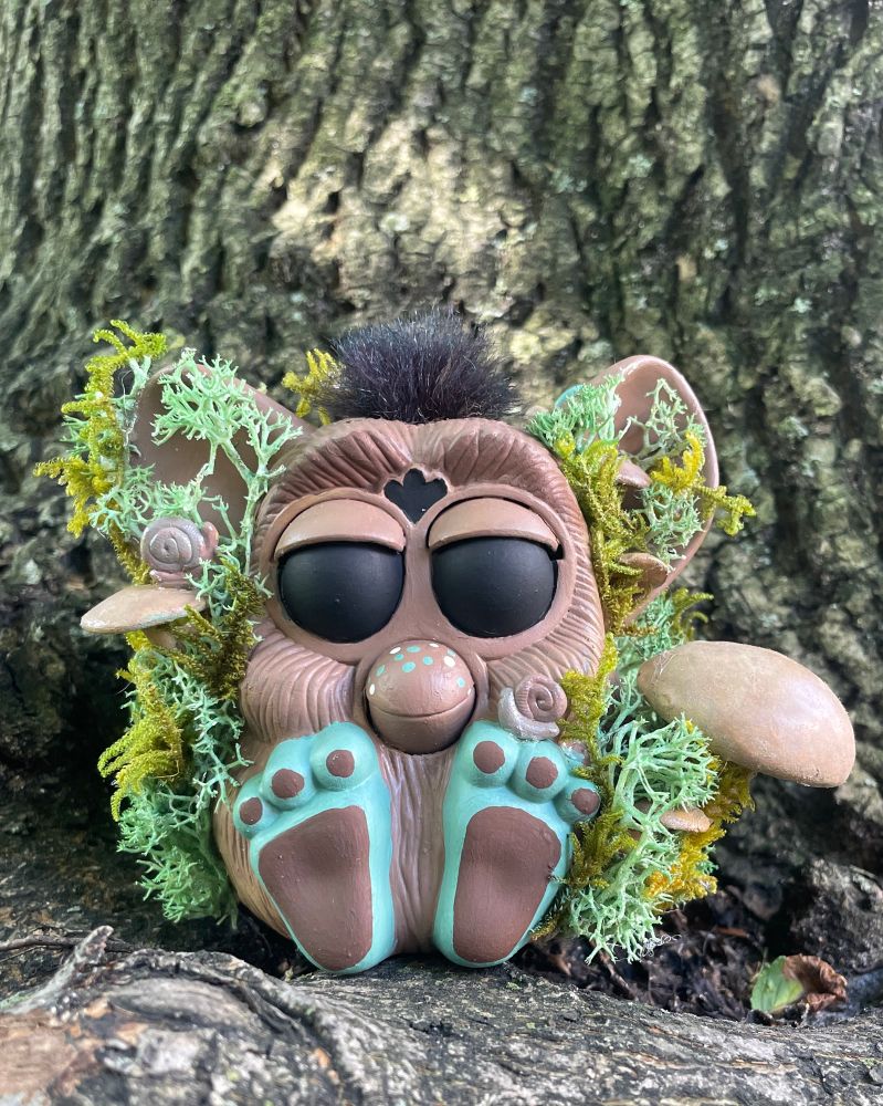 a happy meal furby that’s been custom painted brown with black eyes and green feet. it is covered in moss and mushrooms and there are two snails hanging out on one mushroom and on the Furby’s left foot.