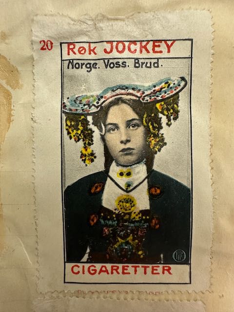 Reklamemerke med fargelagt portrett av kvinne i Vossebunad med brudekrone. Reklameteksten sier Røk JOCKEY CIGARETTER og bildet er påskrevet ordene Norge. Voss. Brud.
