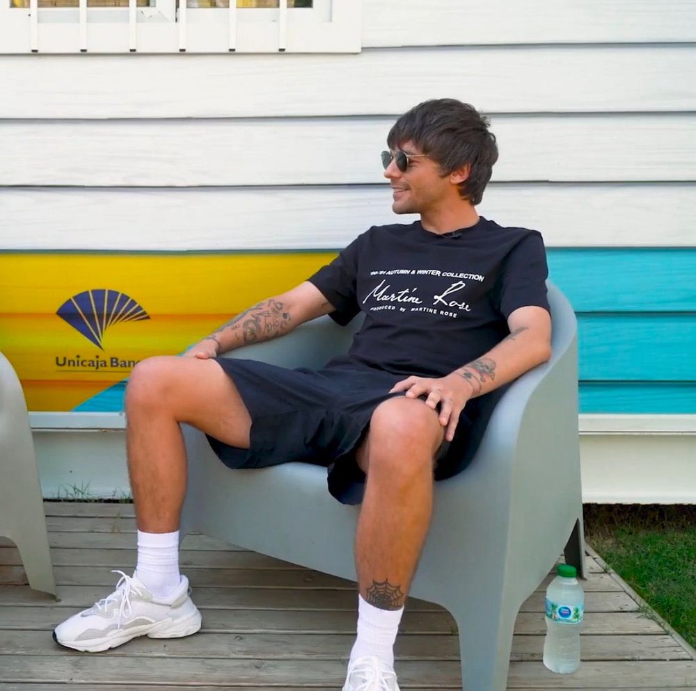 Louis manspreads after the AFHF in Malaga Spain, in black sunglasses. The spiderweb tattoo is visible