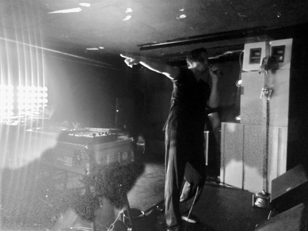 Black and white image of a man on stage, arms outstretched with bright light on the left and speaker stack on the right.